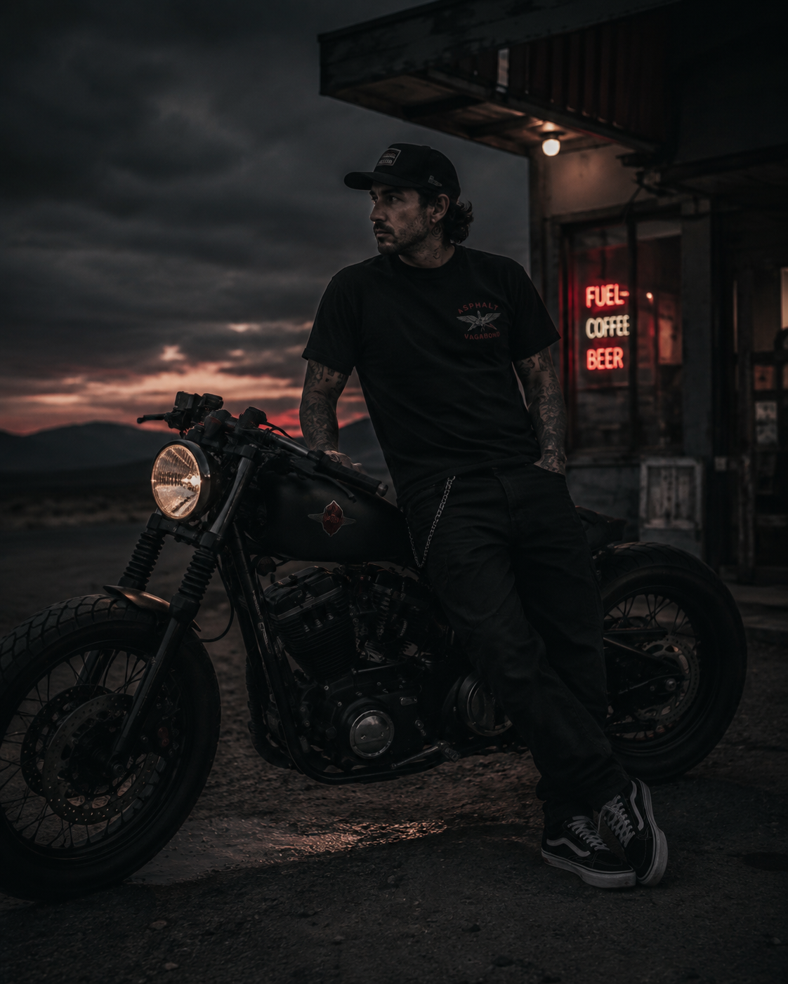 Asphalt Vagabond rider leaning on a custom motorcycle at a roadside fuel stop at dusk