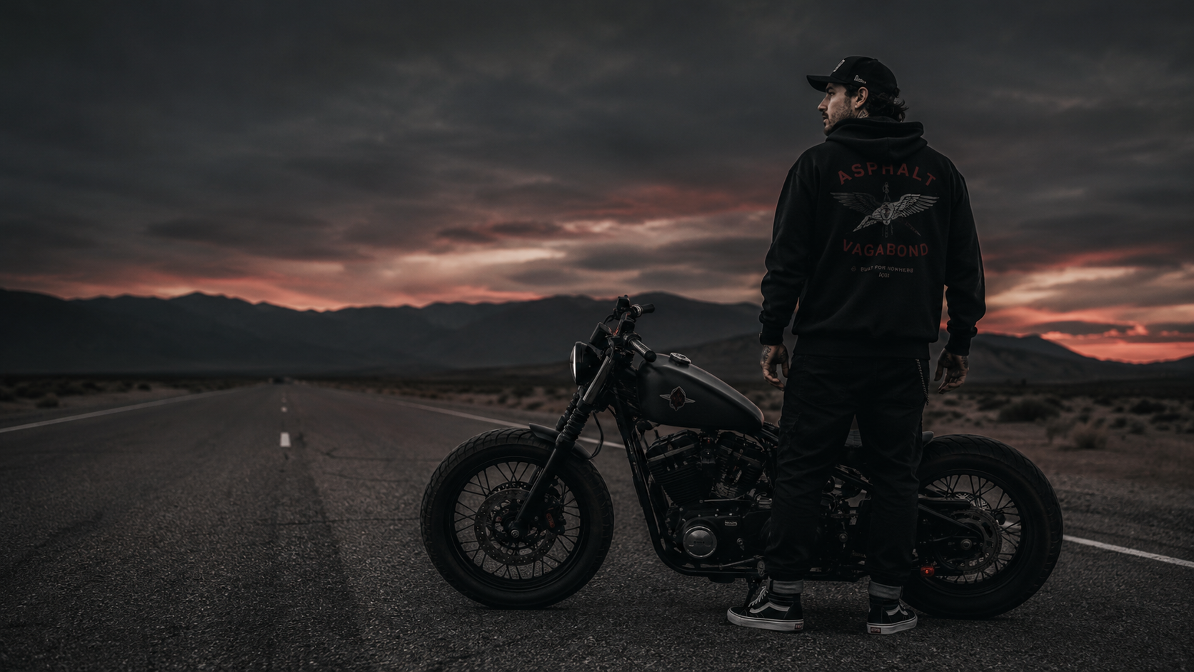 Asphalt Vagabond rider beside a custom motorcycle on an open highway at dusk